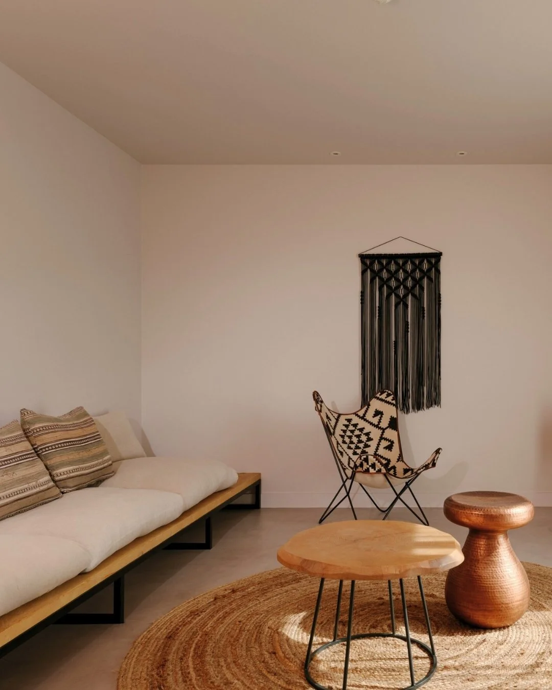 Minimalist living room with a white couch, decorative pillows, a butterfly chair with a patterned cushion, a small wooden coffee table, a rounded wooden side stool, a circular woven rug, and a black wall hanging on a plain white wall.