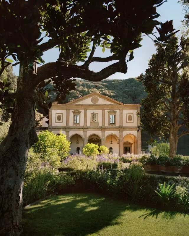 A historic building with classical architecture featuring arches on the ground floor, decorative window frames, and a triangular pediment, situated in a garden with lush green trees, shrubs, and flowering plants, with mountains in the background under a clear sky.