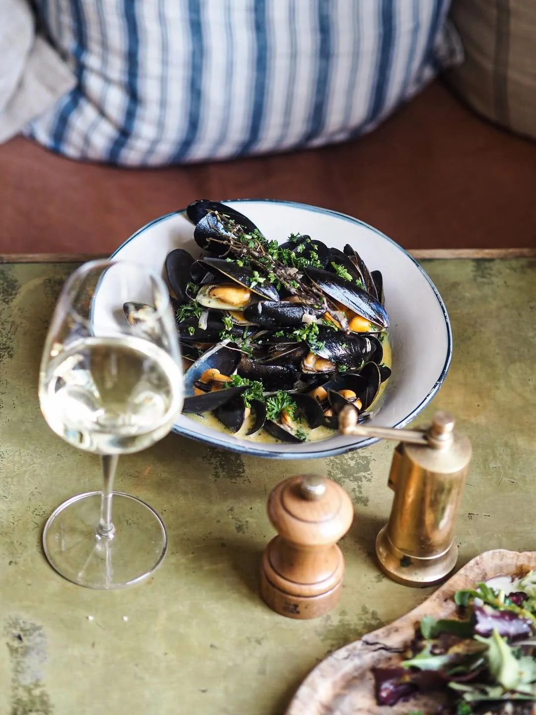 A bowl of cooked mussels garnished with herbs on a green table, beside a glass of white wine and salt and pepper shakers.