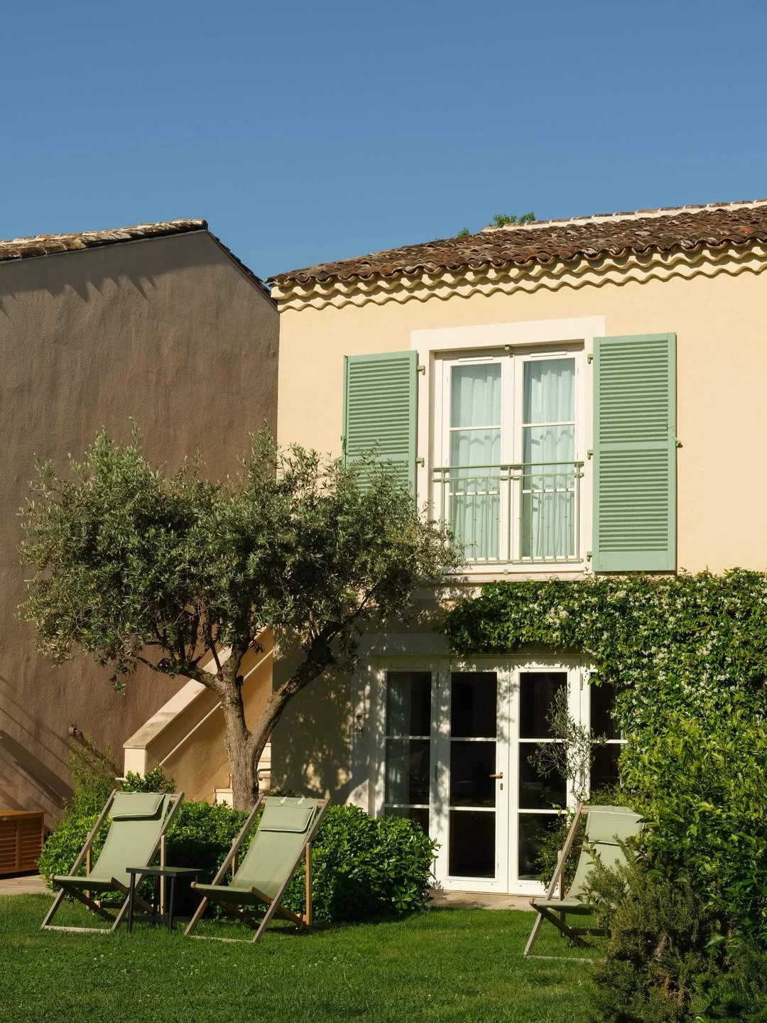 A two-story house with yellow walls, green shutters, and a tile roof. A small balcony with a metal railing is on the second floor. The yard features two lounge chairs, a small table, and various bushes and trees under a clear blue sky.