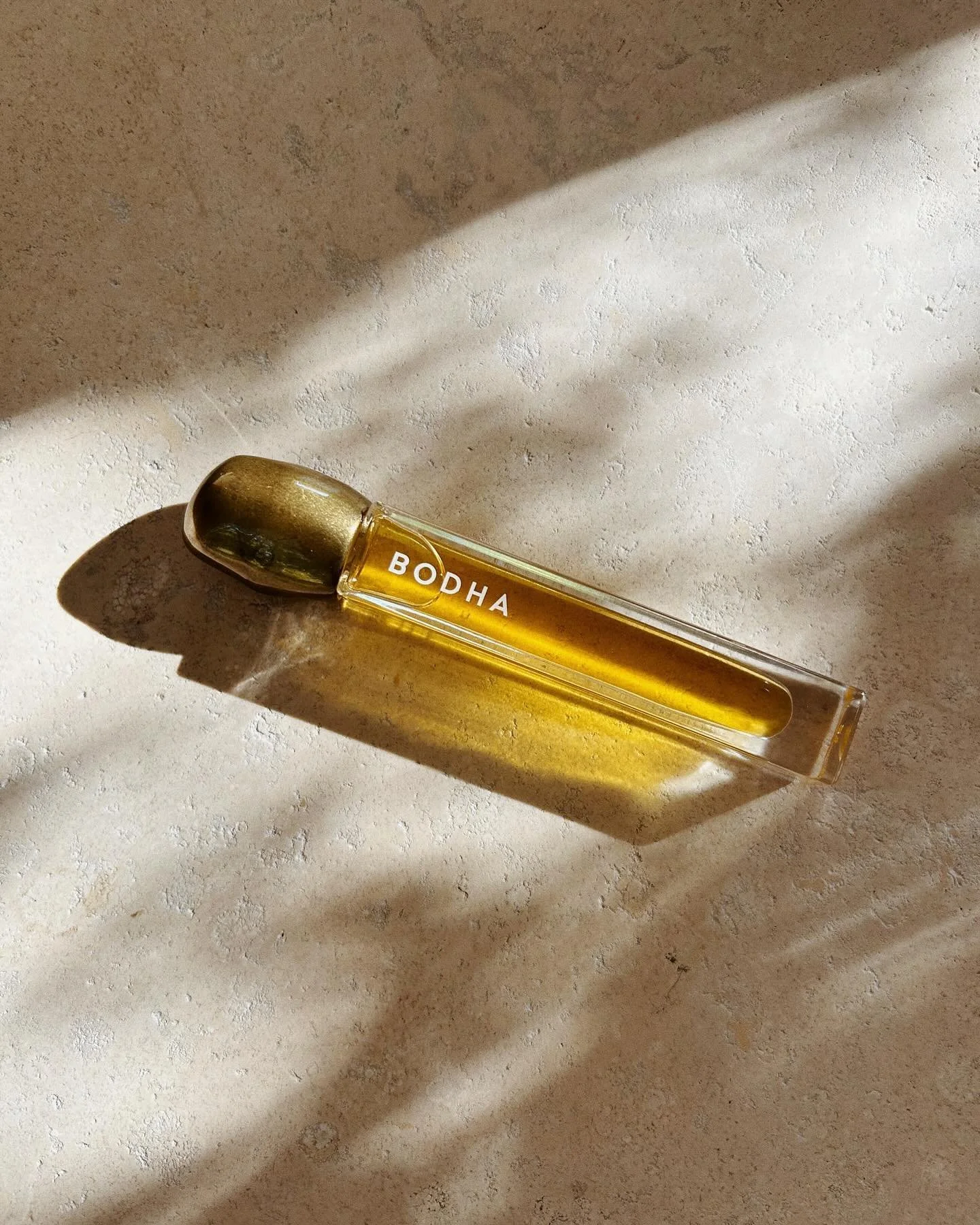 A glass bottle labeled 'BODHA' filled with a yellow liquid, placed on a light-colored textured surface with shadows cast by sunlight.