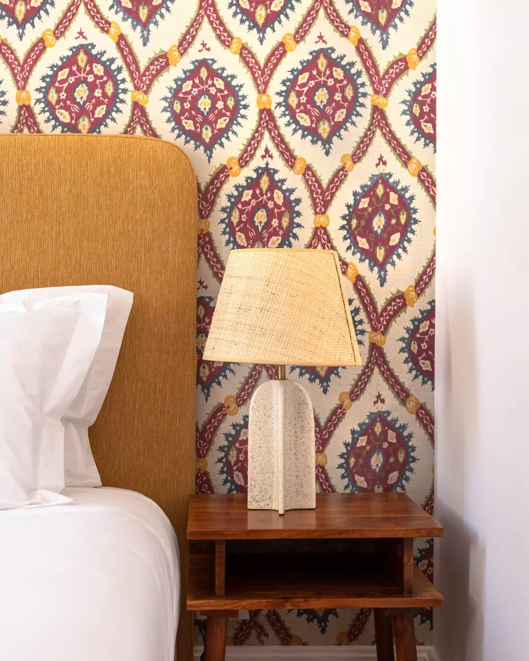 A bedside table with a textured white ceramic lamp with a beige lampshade, placed against a patterned wallpaper with red, blue, yellow, and cream geometric and floral designs, adjacent to a bed with a mustard-colored headboard and white bedding.