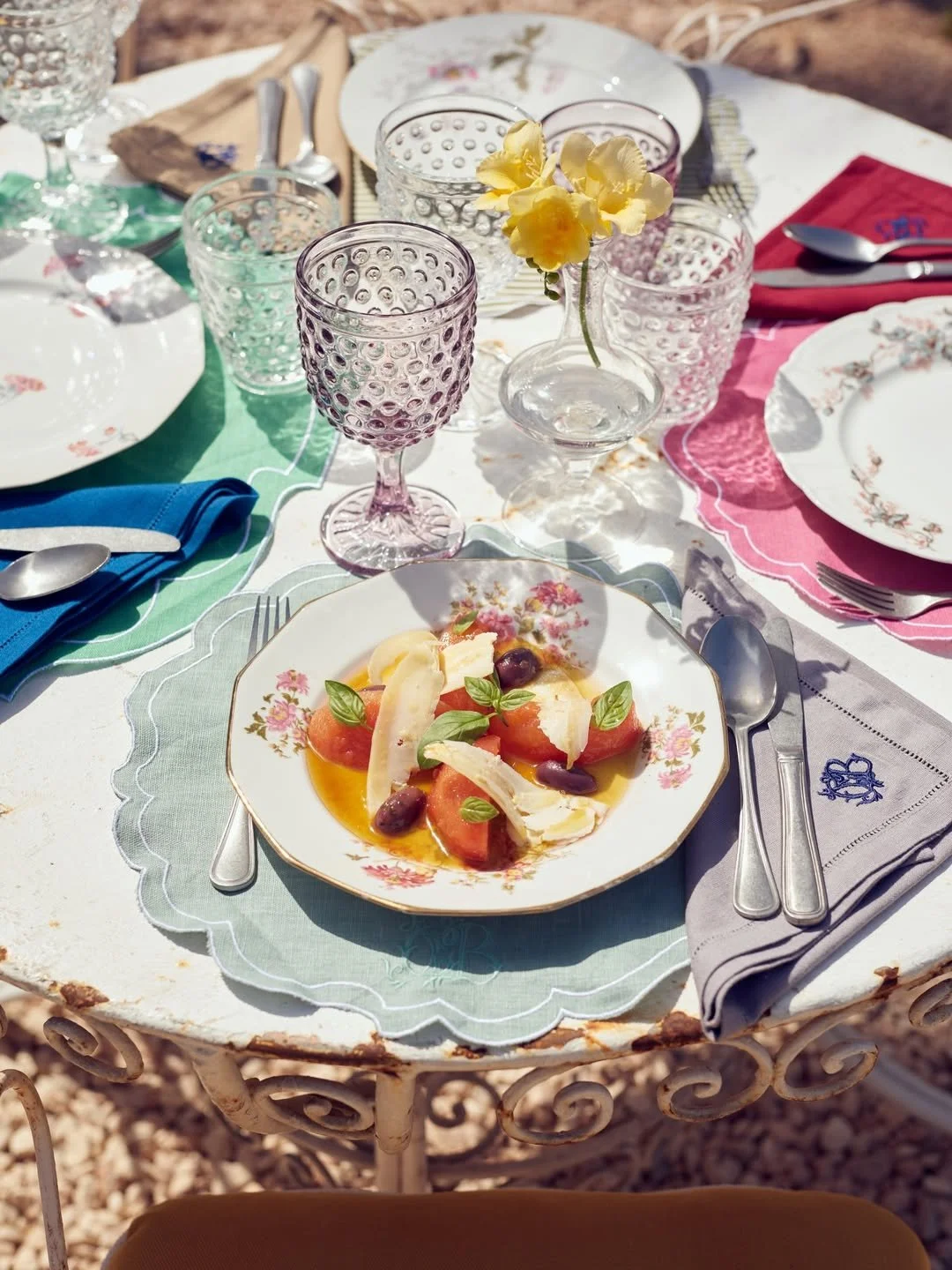 Elegant table setting with floral china plates, colorful glasses, silverware, and a plate of watermelon with cheese, olives, and basil.