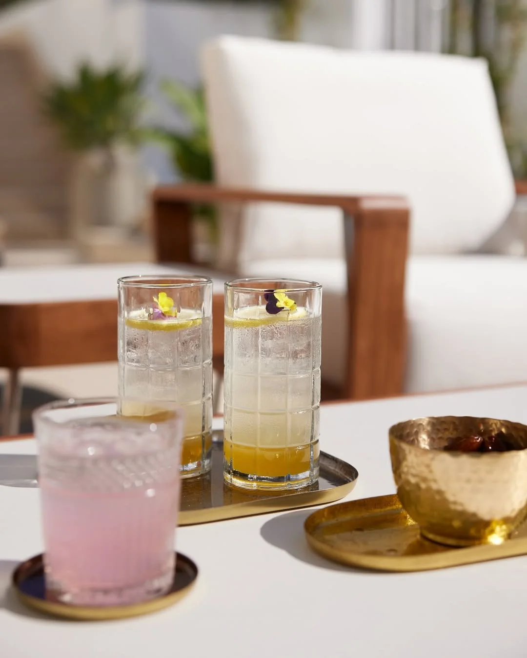 Two tall glasses with yellow beverages garnished with purple and yellow edible flowers, a pink drink in a shorter glass, and a small gold bowl on a white table with gold trays.