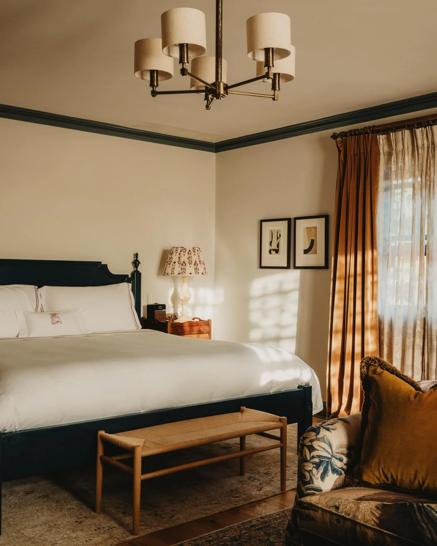 A cozy bedroom with a large bed, white bedding, a wooden bench at the foot of the bed, a patterned table lamp on a nightstand, framed artwork on the wall, a window with gold curtains, and a vintage armchair with a gold pillow, all illuminated by warm natural light.