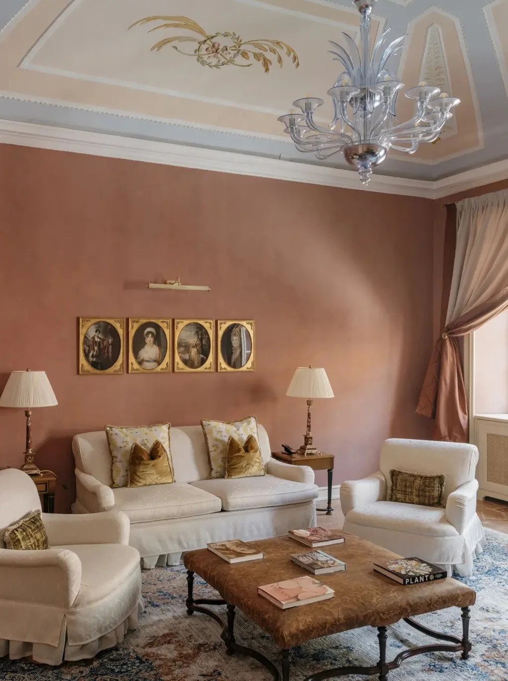 Elegant living room with a peach-colored wall, vintage-style white sofas and armchairs with plush pillows, a wooden coffee table with books, standing lamps, classic framed portraits on the wall, a large window with draped curtains, and a decorative ceiling with a chandelier.