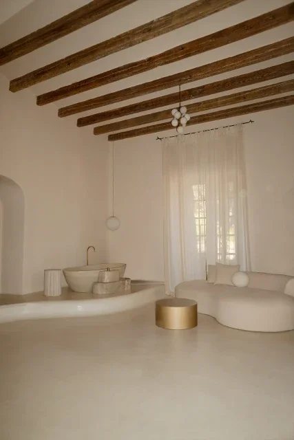 Minimalist living room with white curved furniture, a white curtain in front of a window, and exposed wooden ceiling beams.