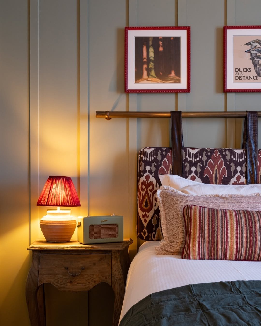 A cozy bedroom corner featuring a bedside table with a red lampshade, a vintage radio, an upholstered bed with patterned headboard, striped pillows, and two framed pictures on the wall.