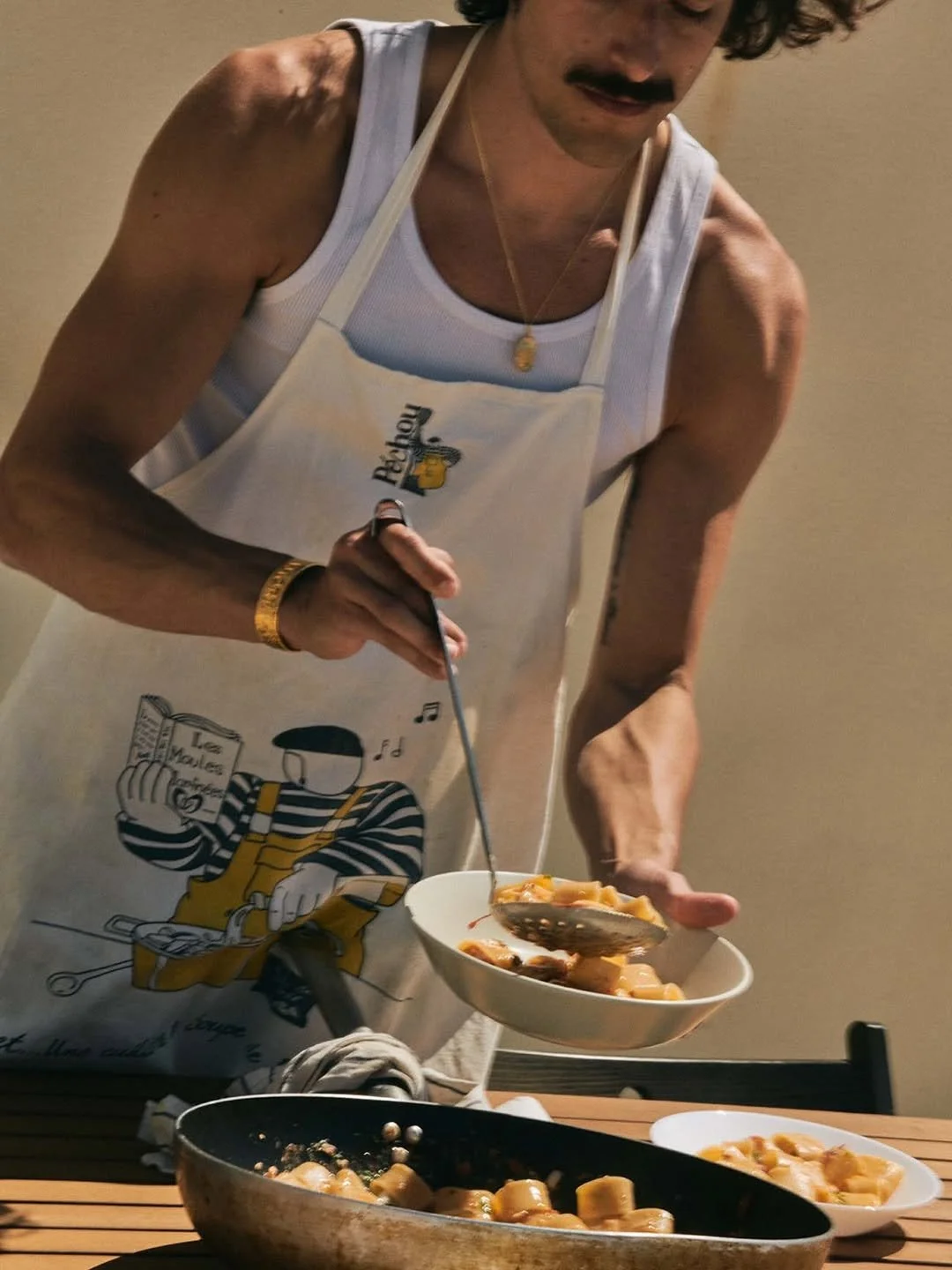 A man with curly hair, wearing a white tank top, a white apron with a cartoon character, and a gold necklace, serving food into a bowl.
