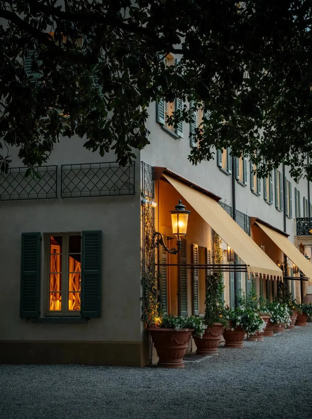 A charming building with multiple windows, green shutters, and a balcony under a tree, illuminated by warm outdoor lights and lamps, with potted plants along the exterior.
