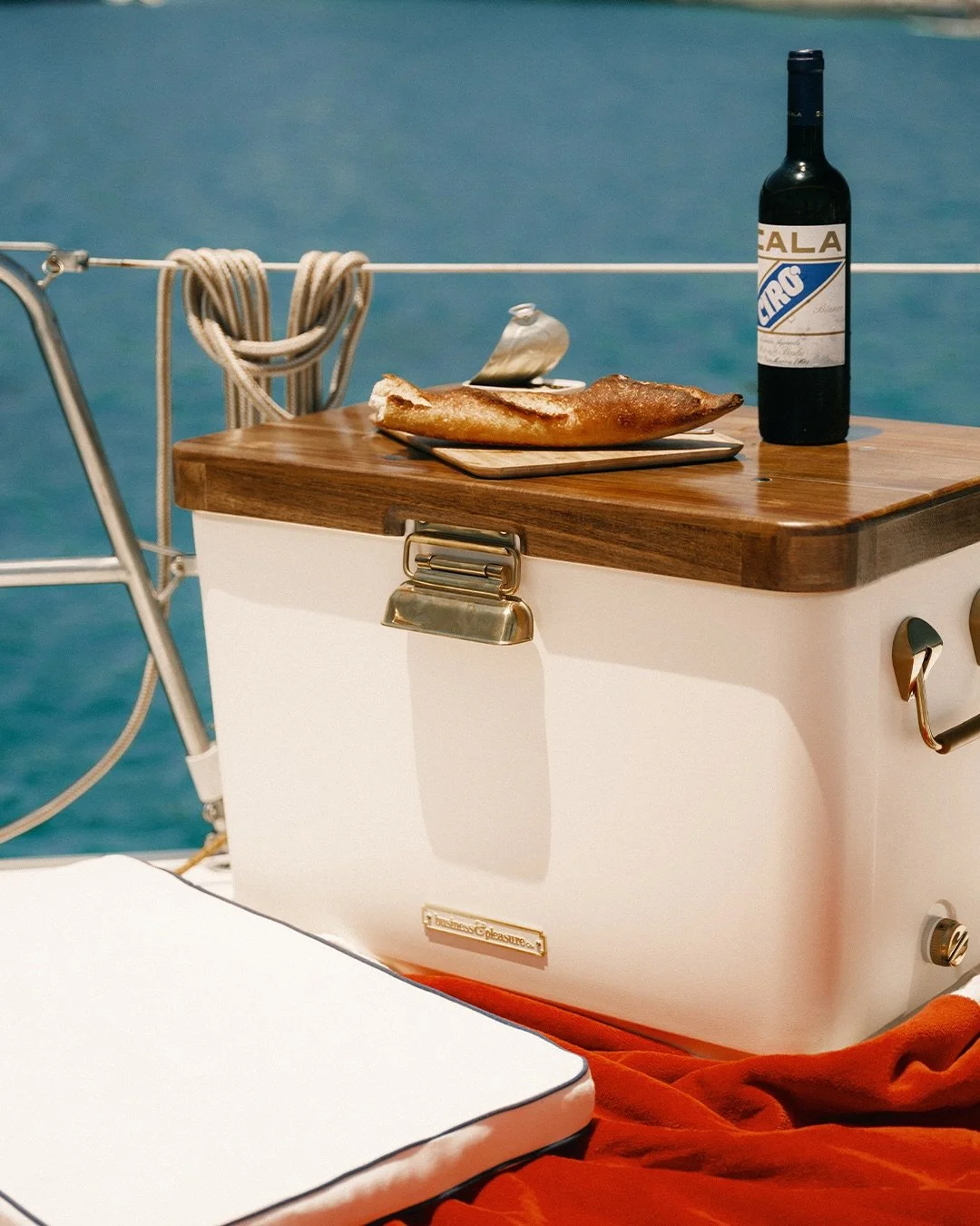 A wooden table on a boat with a bottle of red wine, a plate with a piece of bread or pizza, and a blue seas background.