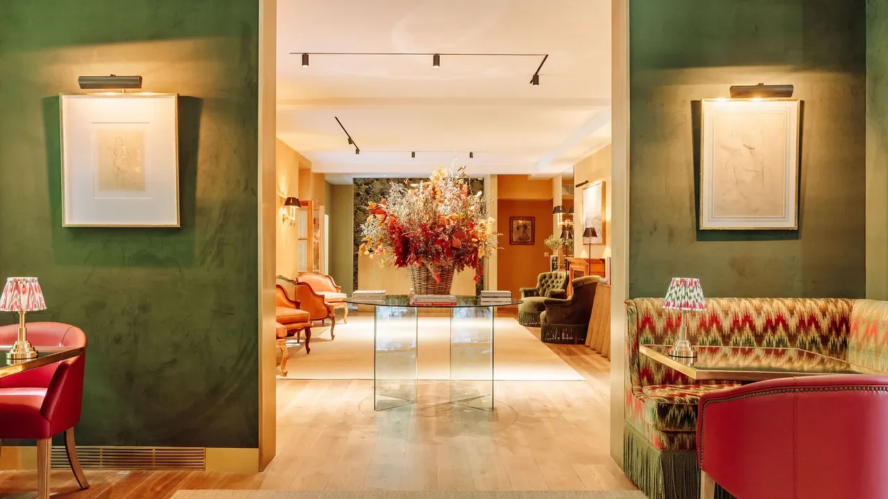A stylish hotel lobby with green walls, framed artwork, and colorful furniture including a patterned booth and pink chairs. A large floral arrangement is centered on a glass table, with additional seating and artwork visible in the background.