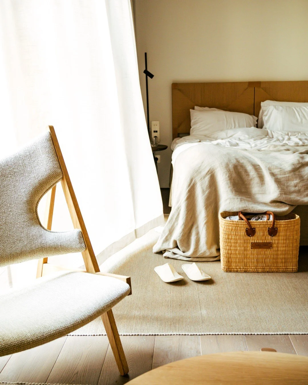 A neatly made bed with white pillows and bedding, a wicker basket in front of the bed, a pair of white slippers on the beige carpet, a wooden chair with a light-colored cushion on the left, and a small bedside table with a dark lamp behind the bed, in a well-lit room with curtains.