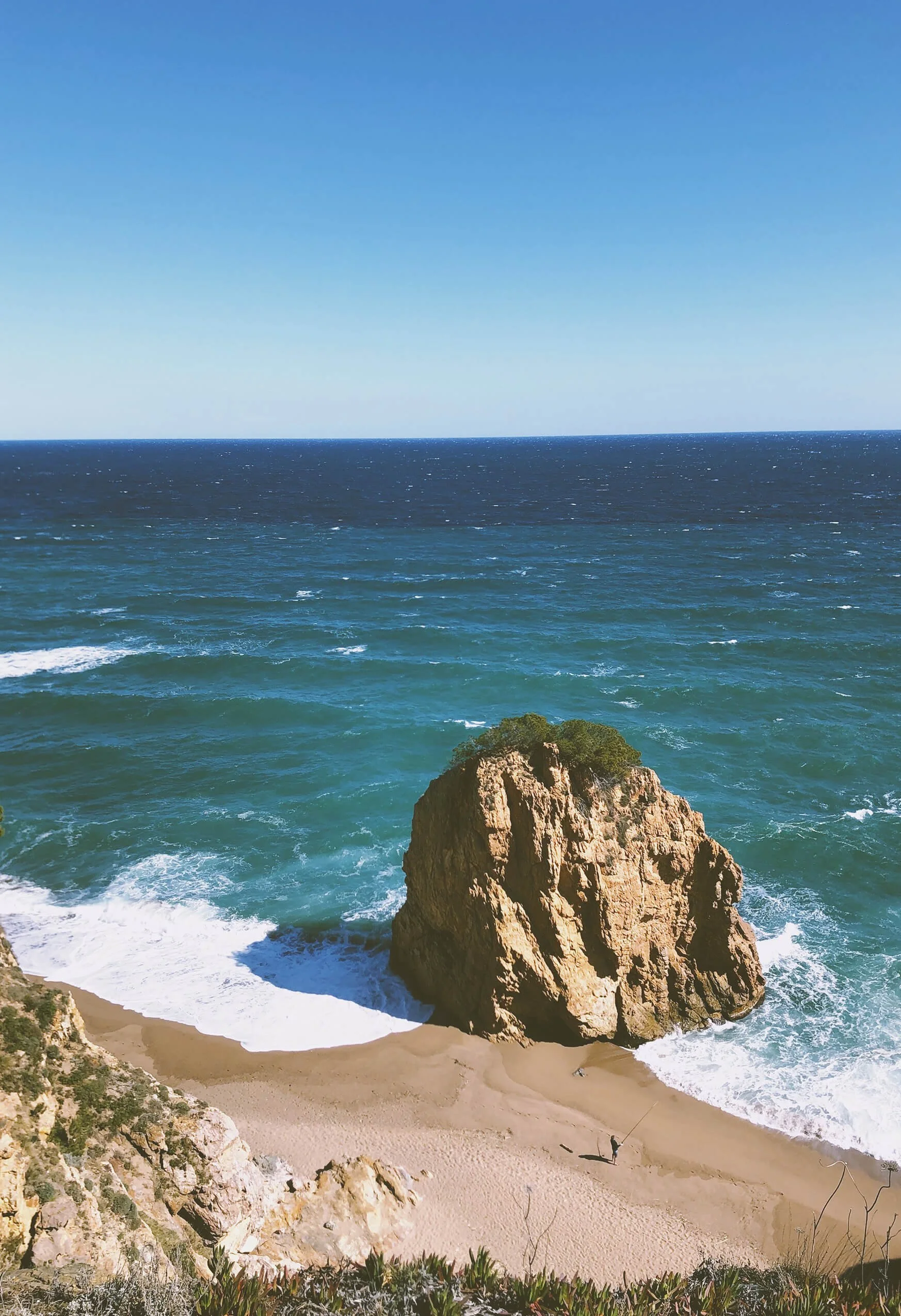 A scenic coastal view with a large rock formation on the beach, waves crashing against it, clear blue sky, and a person fishing on the sand.