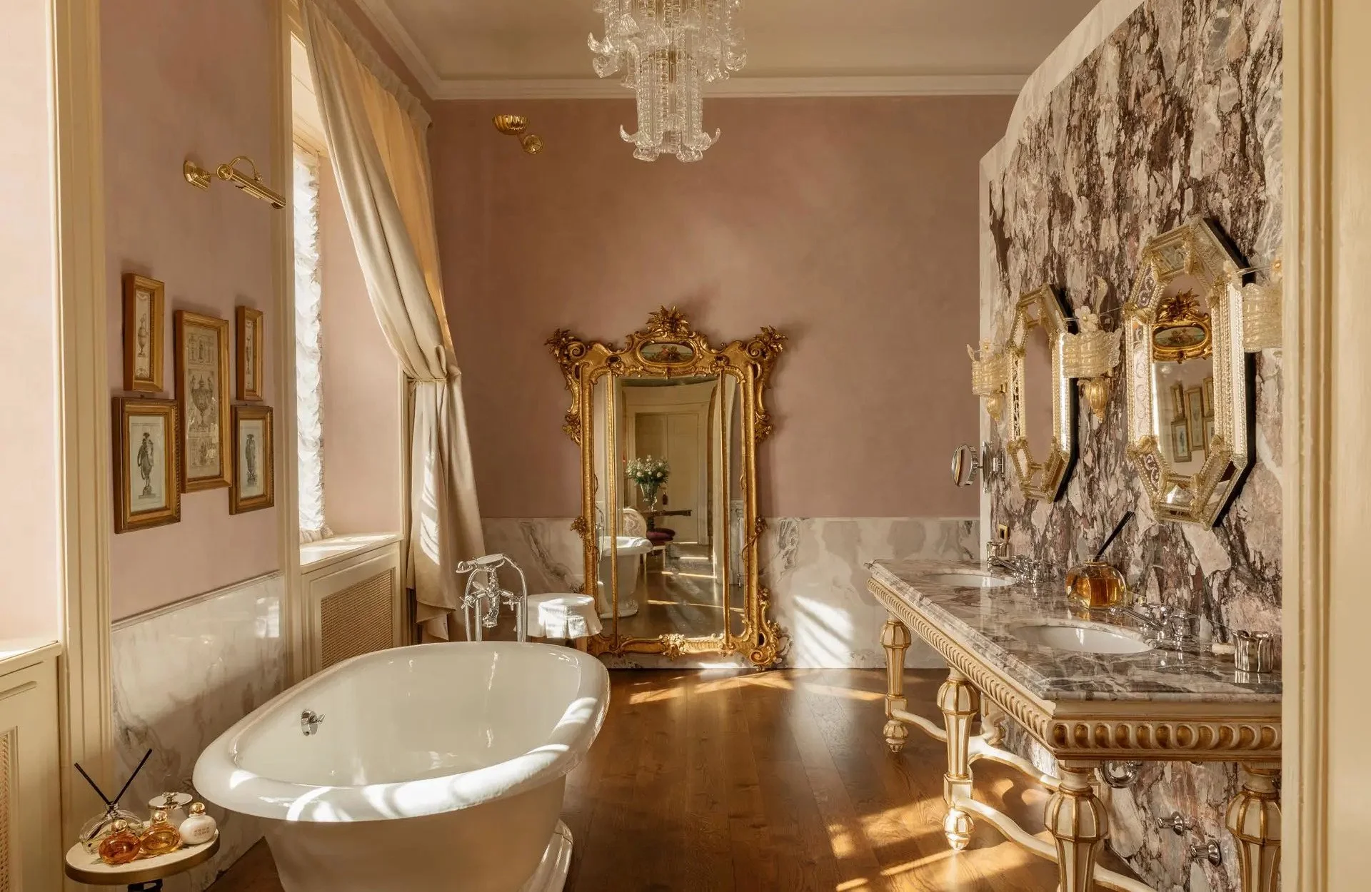 Elegant bathroom with a vintage bathtub, ornate gold mirror, marble countertop, framed artwork, and chandelier in a classical style.