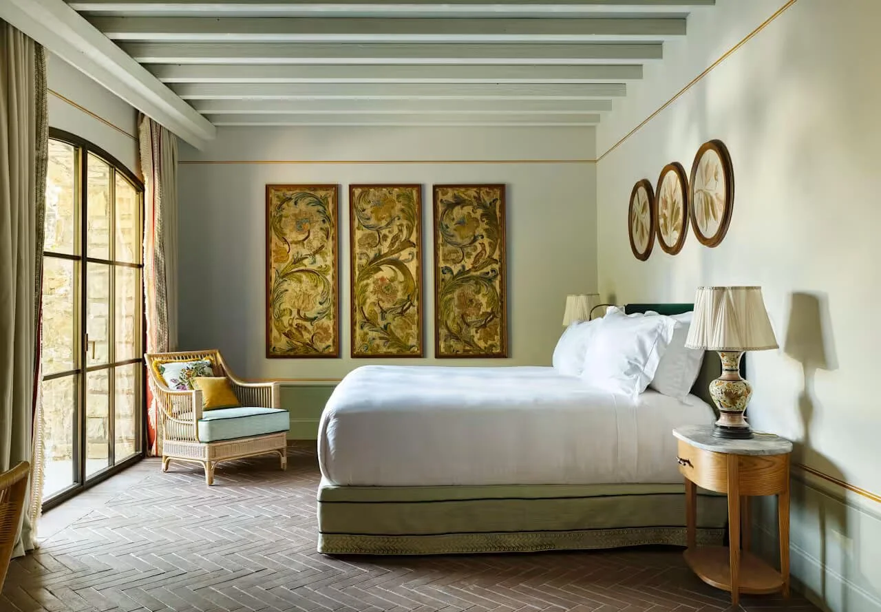 A bedroom with a large bed, nightstand with a lamp, armchair with a cushion near window, decorative wall art, and sunlight streaming through glass doors.