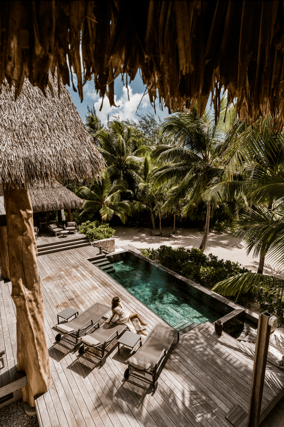 A woman relaxing on a lounge chair by a small pool on a wooden deck surrounded by tropical trees, with thatched roof structures and sandy beach nearby.