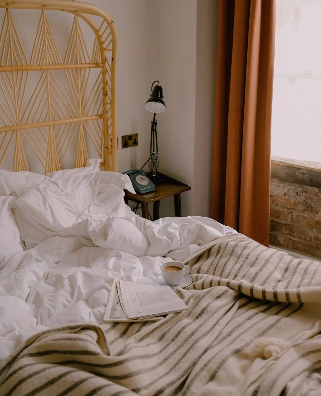 Unmade bed with white sheets and a striped blanket, a cup of coffee, an open newspaper, a vintage phone, a black desk lamp, and a window with brick wall outside, in a cozy bedroom.