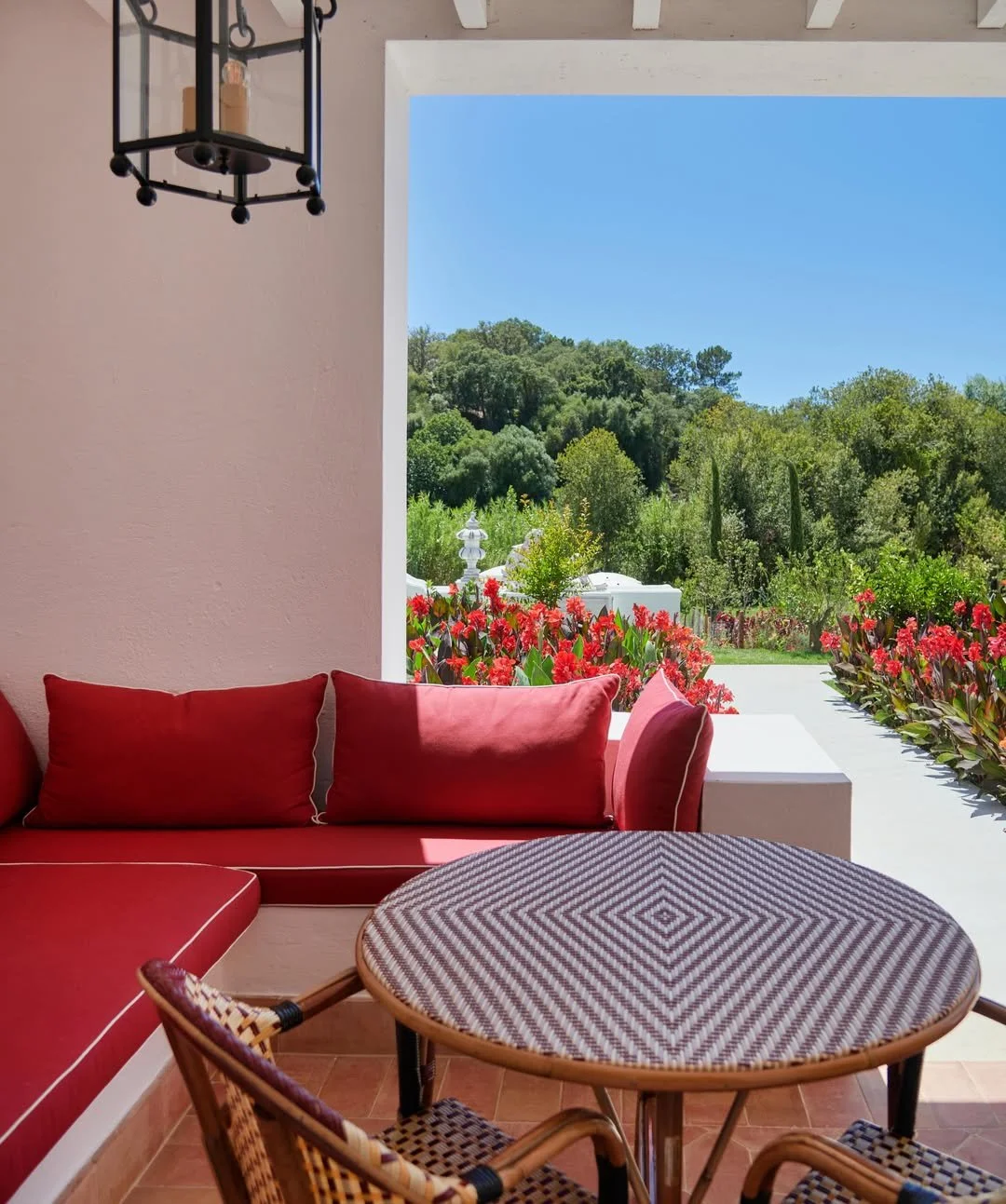 A porch or patio view with a red cushioned sofa, a round woven table, and two woven chairs; outside, red flowers and lush green trees under a clear blue sky.