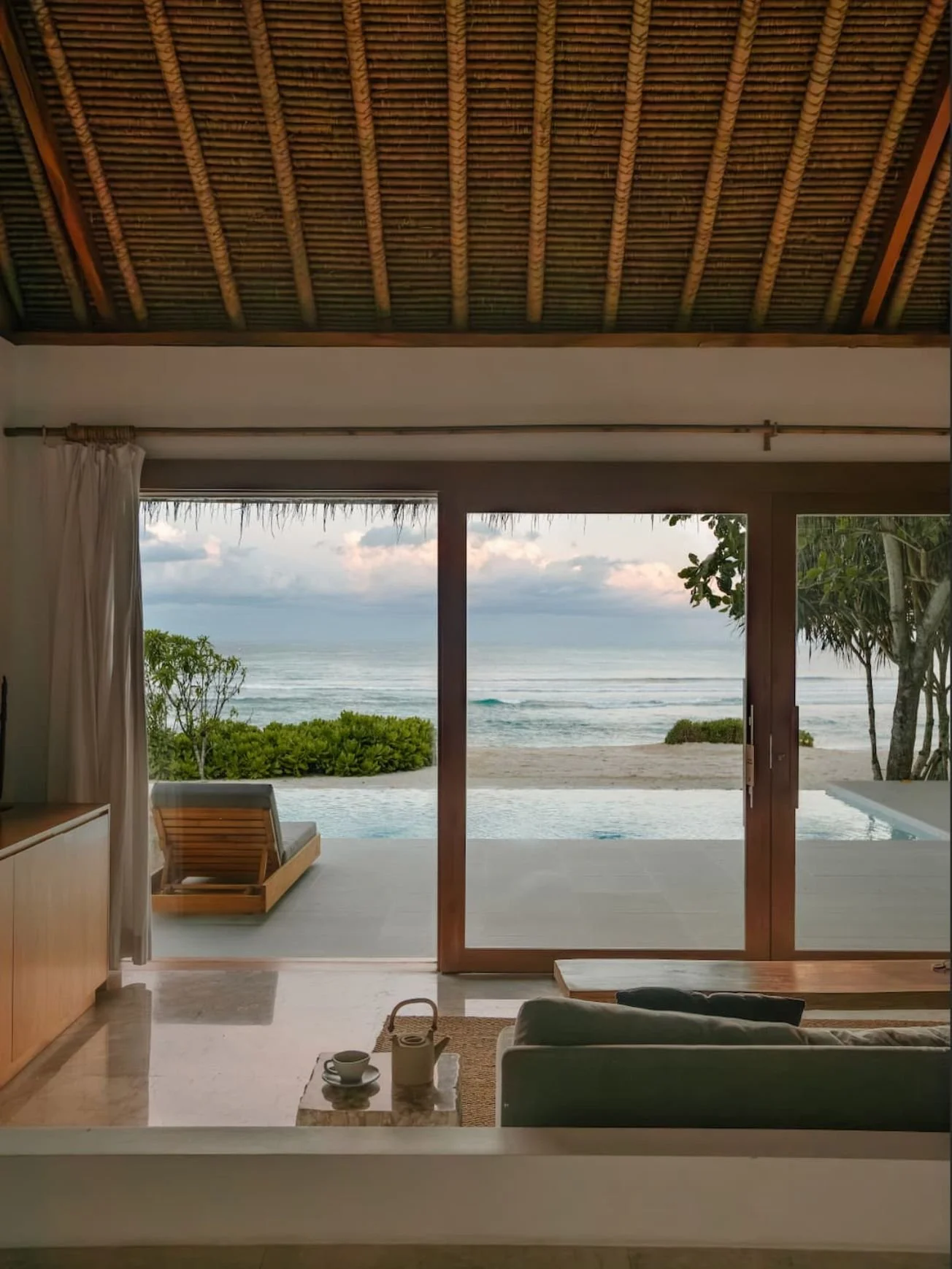 View of a tropical beach from a room with sliding glass doors, featuring an outdoor pool, lush greenery, and ocean waves under a partly cloudy sky.
