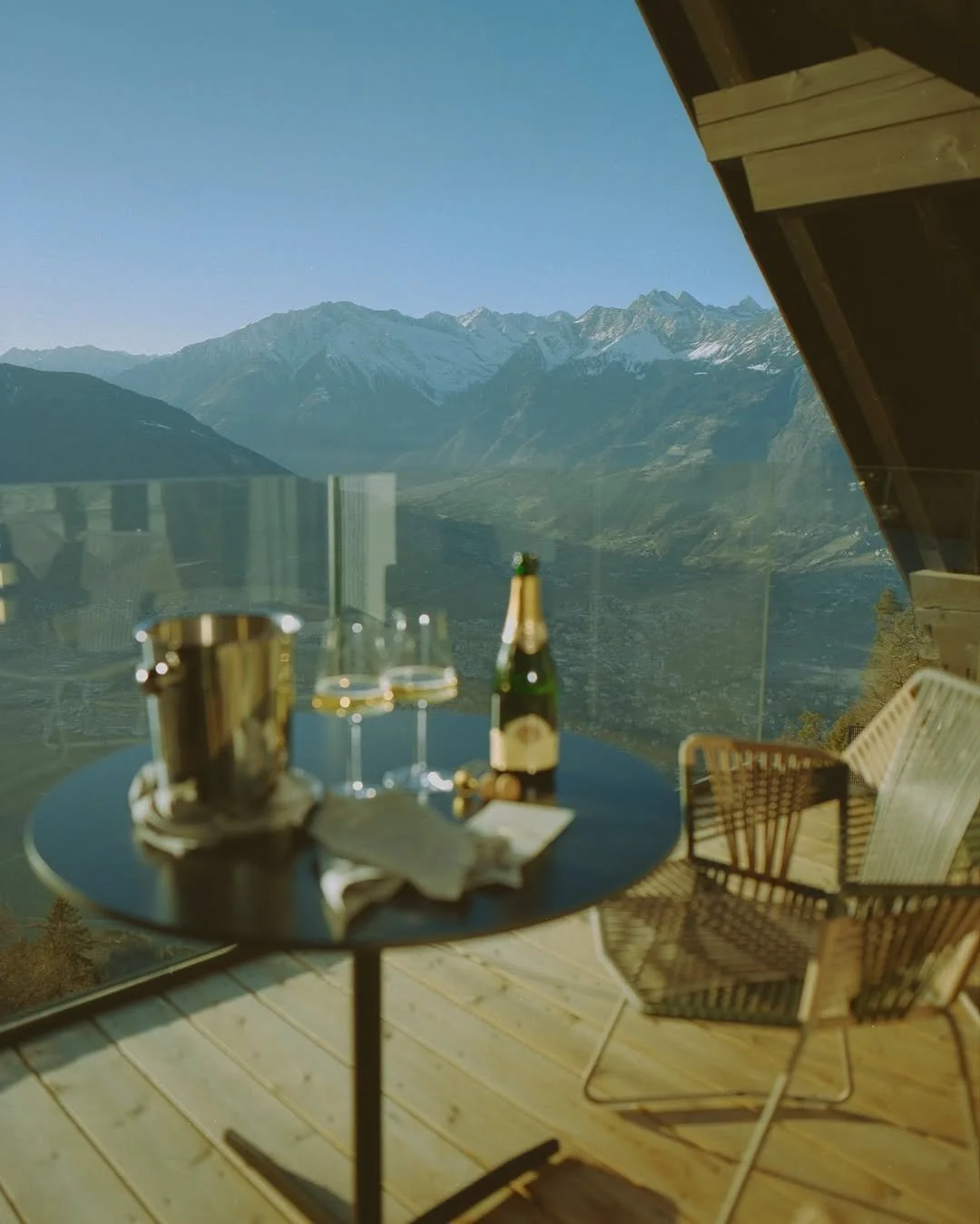 Table with champagne bottle, glasses, and ice bucket on a balcony overlooking mountains and a valley.