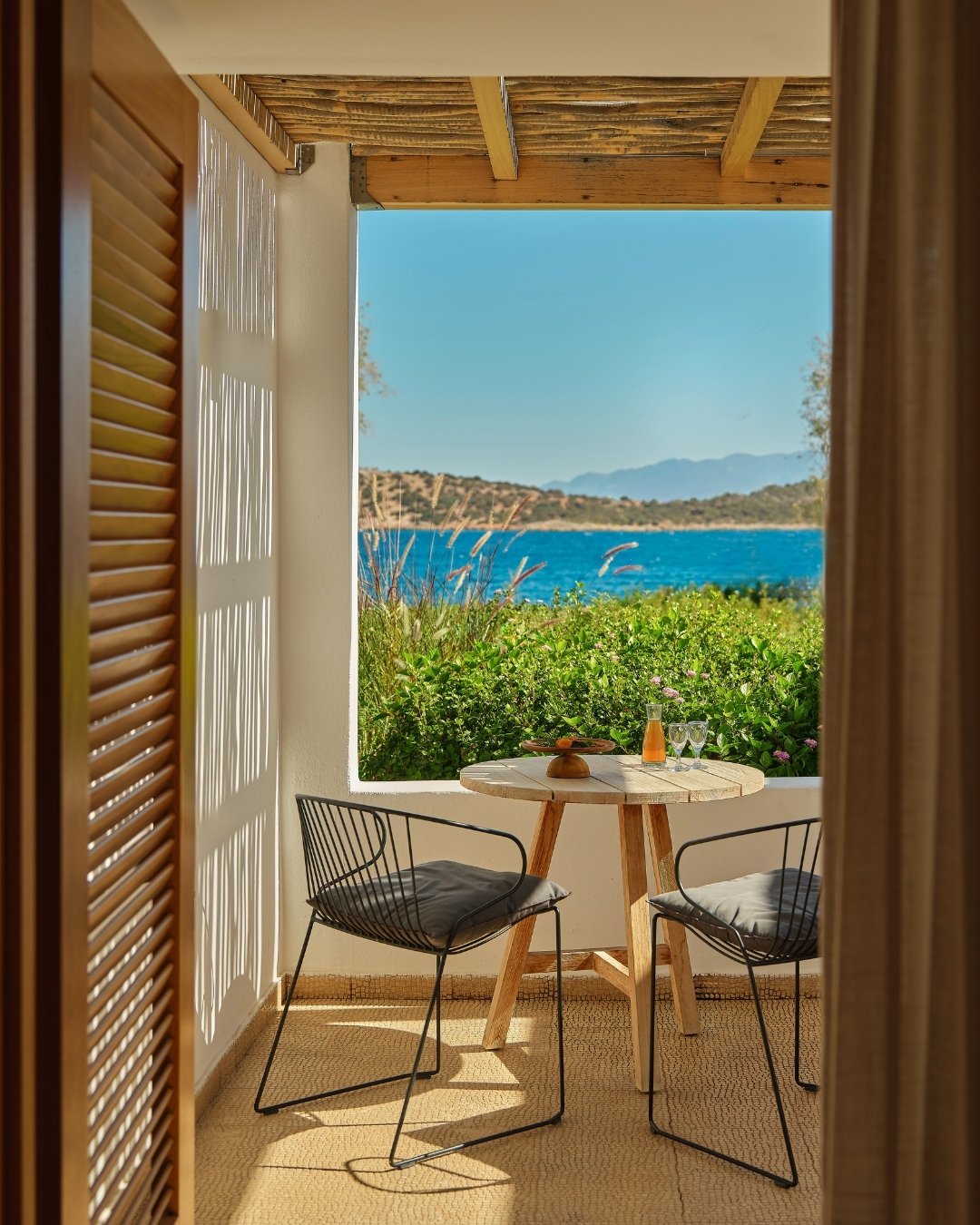 View from a cozy outdoor patio with a wooden table set for two, overlooking a blue lake and distant mountains under clear sky.
