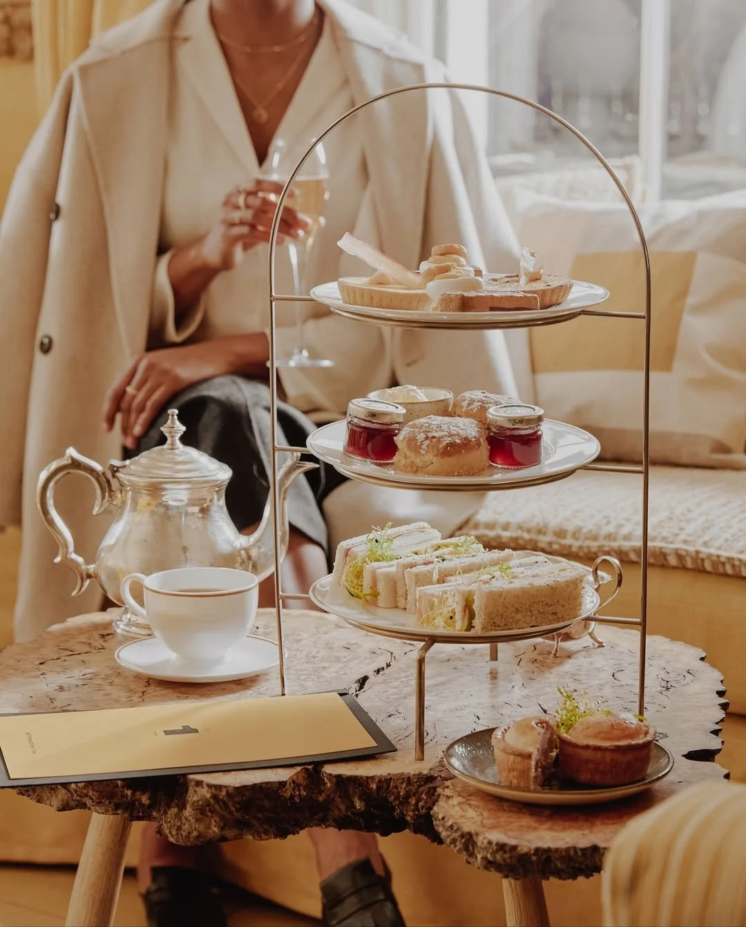 Three-tiered stand with assorted pastries and finger sandwiches set on a rustic wooden table, with a teapot, teacup, and a woman in the background holding a glass of tea in a cozy, well-lit room.