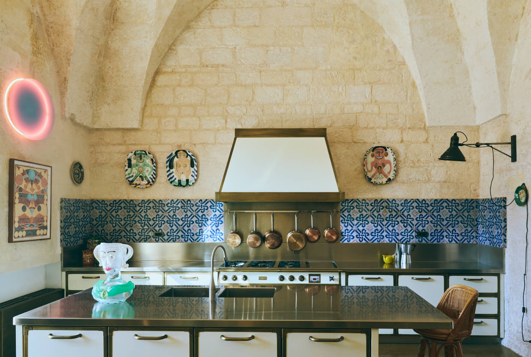 Modern kitchen with a stainless steel island, hanging copper pots, decorative ceramics on the wall, and an arched stone ceiling.