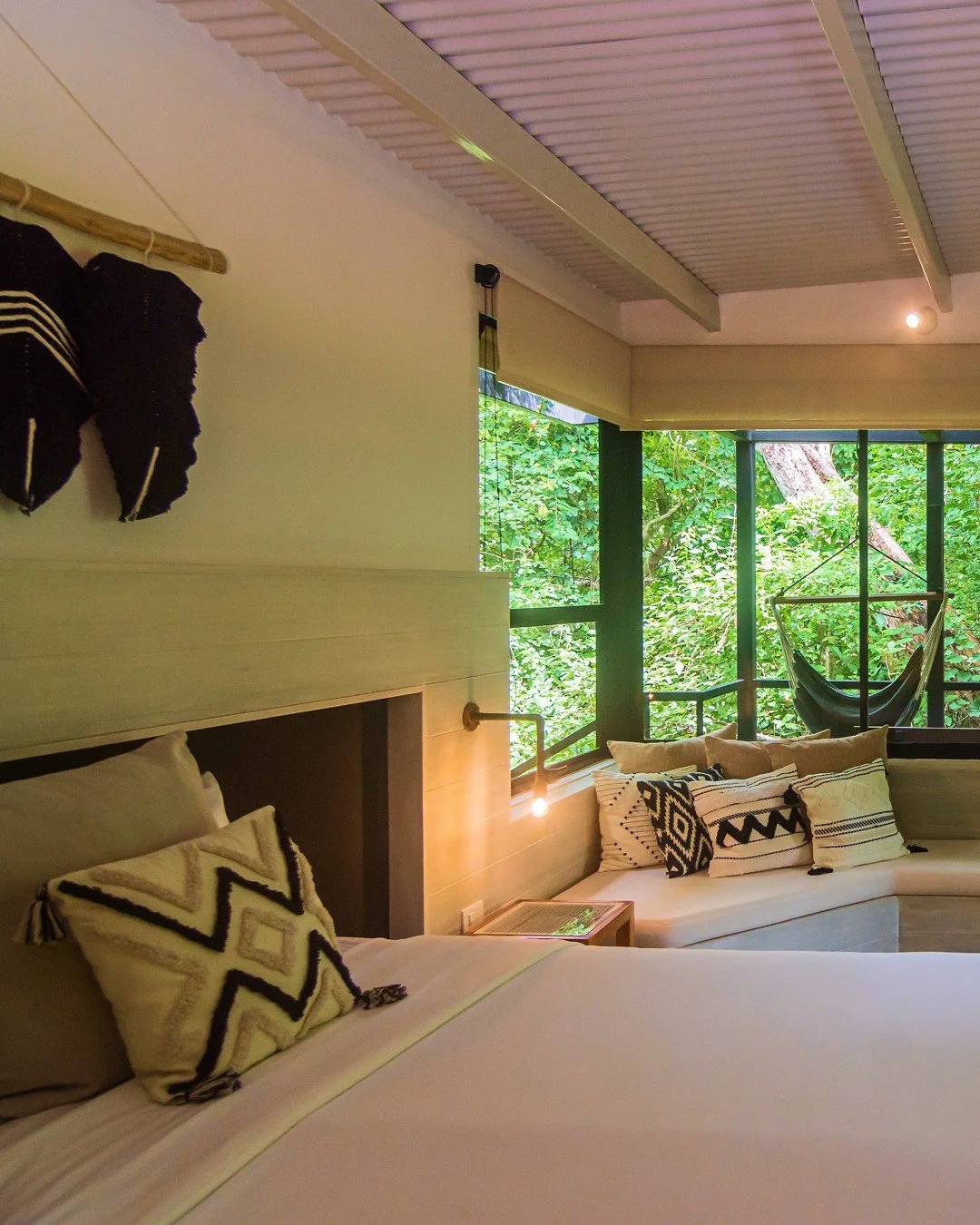 A cozy hotel room with a bed, decorative pillows, a small side table with a glass, large windows showing lush trees outside, and a hammock hanging near the window.