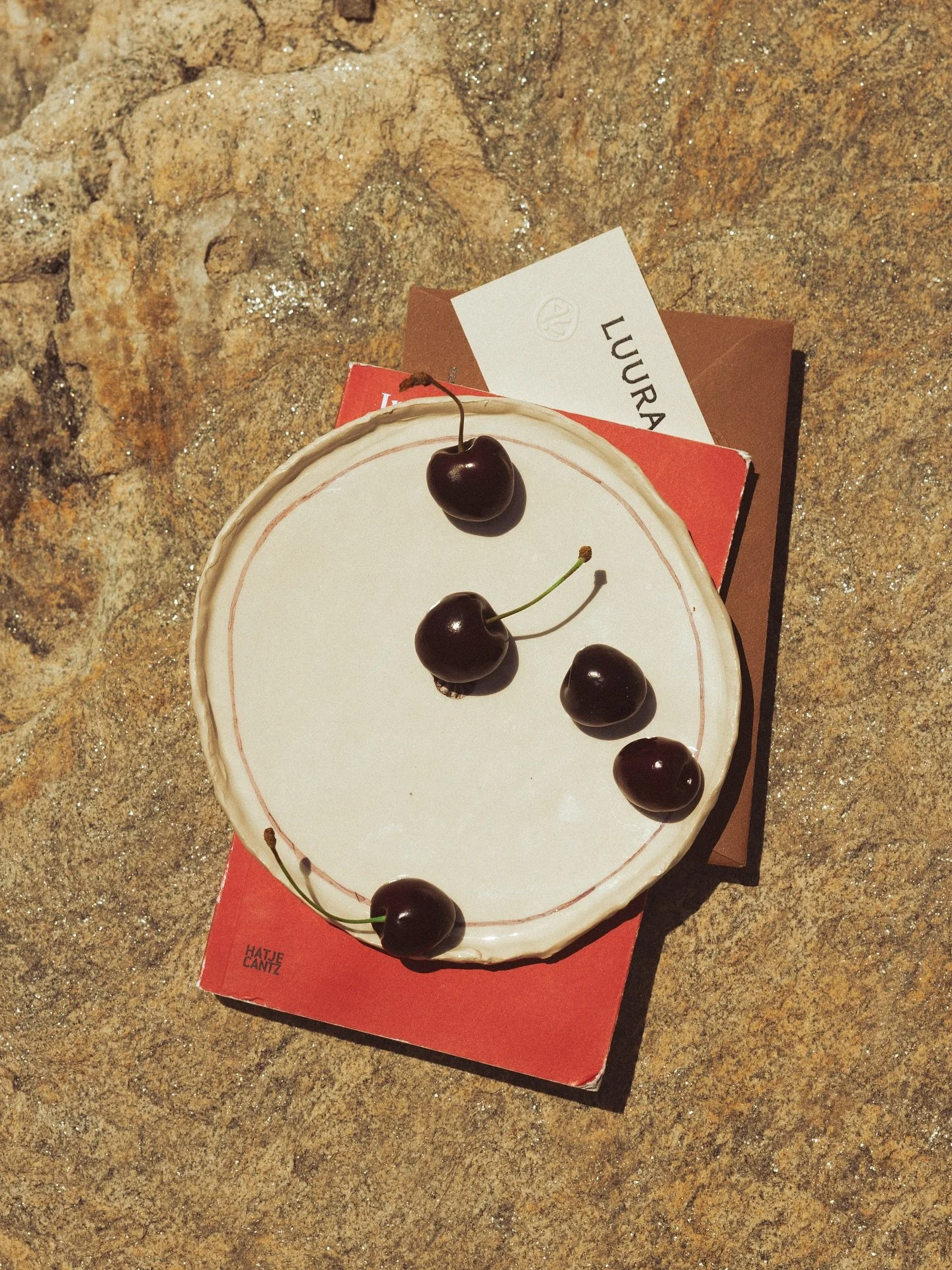 A round white cake with a thin pink border, topped with five dark cherries. The cake is on a red box with text and a white business card behind it, all placed on a rough, sandy surface.