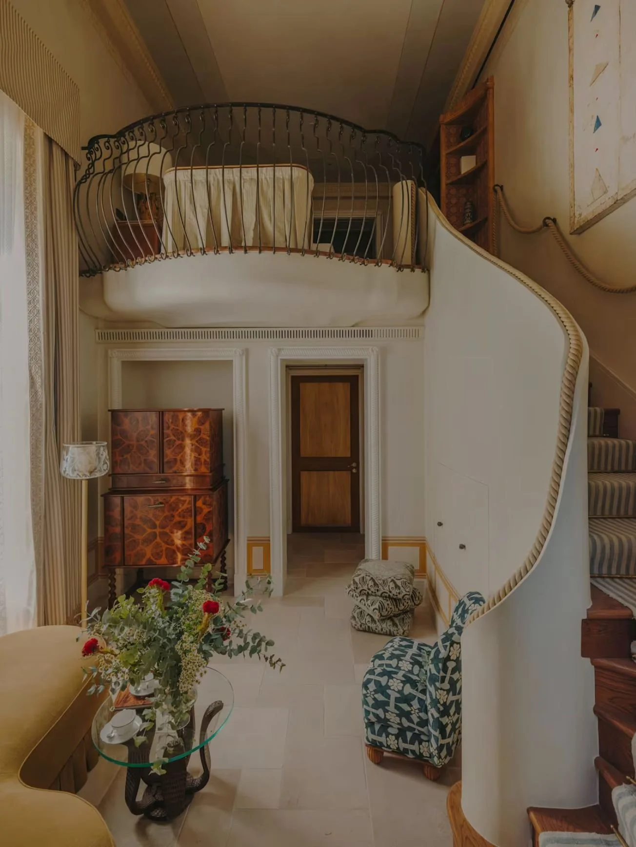 Interior view of a home with a staircase, a small balcony, a door, a floral arrangement, a vintage-style wooden cabinet, and seating areas with patterned cushions.