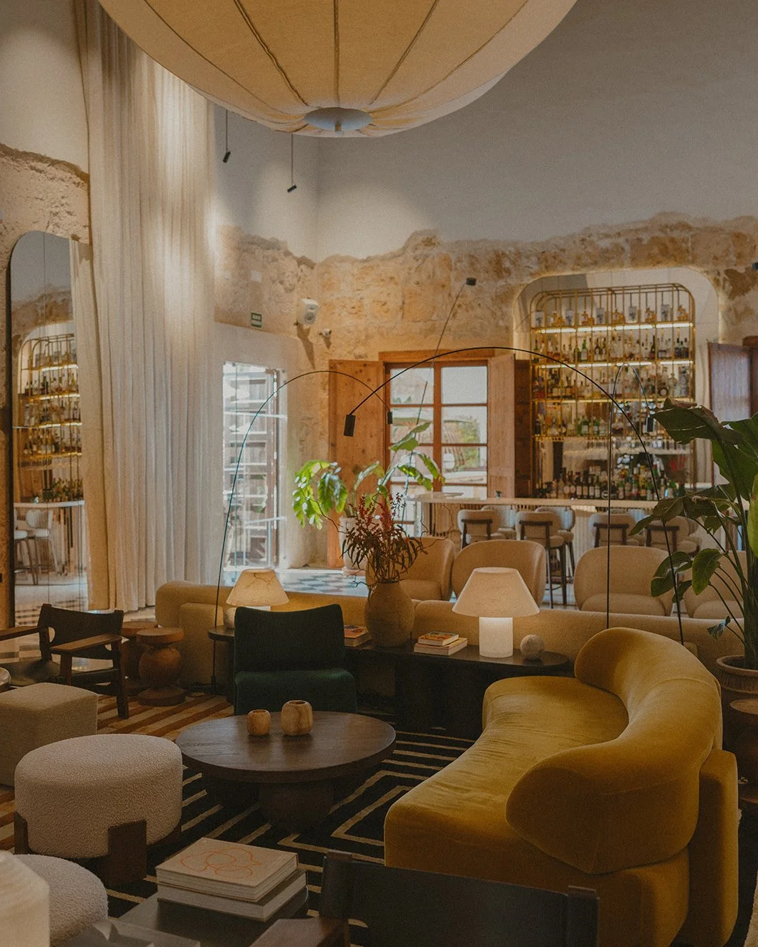 Cozy lounge area with yellow and beige furniture, a black coffee table with books and candles, potted plants, lamps, and a bar area in the background with bottles on shelves, exposed stone walls, and large windows.