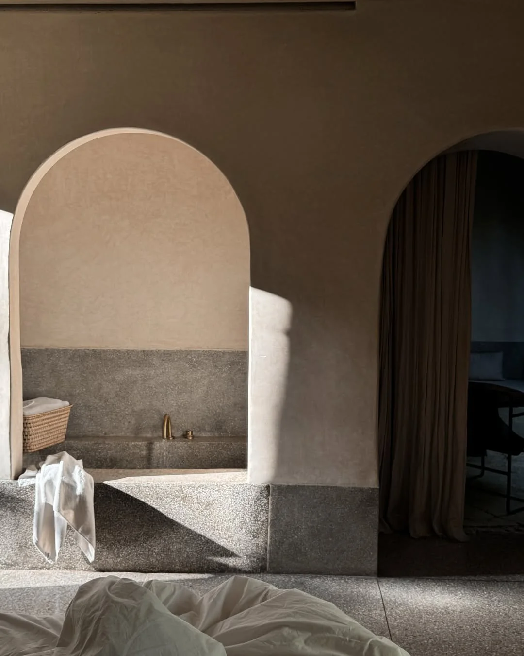 A sunlit bathroom with a stone bathtub, a wicker basket with towels, and a white towel hanging from the edge, with a beige arched wall and a curtain leading to a darker room.
