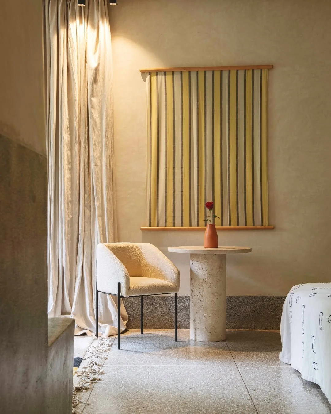 A cozy corner of a room with a beige armchair, a small round table with a red flower in a terracotta vase, beige curtains, and a wall decoration with yellow and gray vertical stripes.