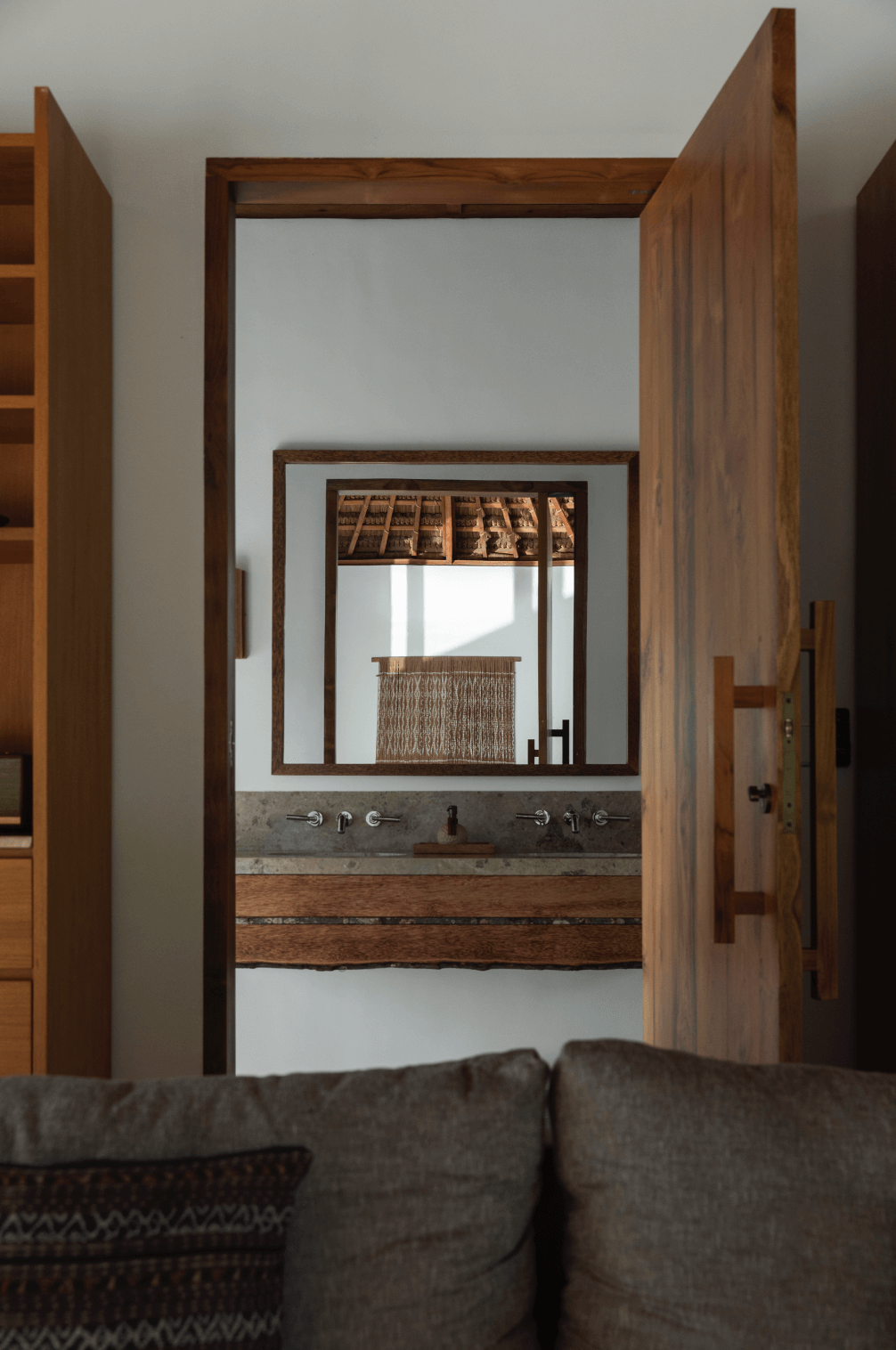 View through an open wooden door into a bathroom with twin sinks, a large mirror, and a sloped ceiling with wooden beams. A window with a curtain is reflected in the mirror.