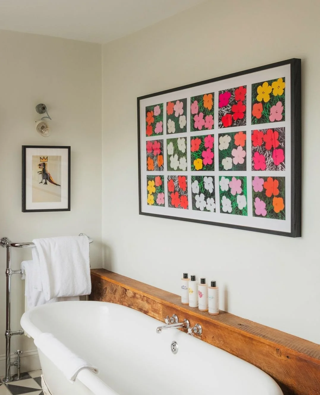 A bathroom with a white clawfoot bathtub, a wooden rail around the tub, five bottles on the rail, and framed artwork on the wall, including a large colorful floral print and a smaller cartoon dinosaur print.