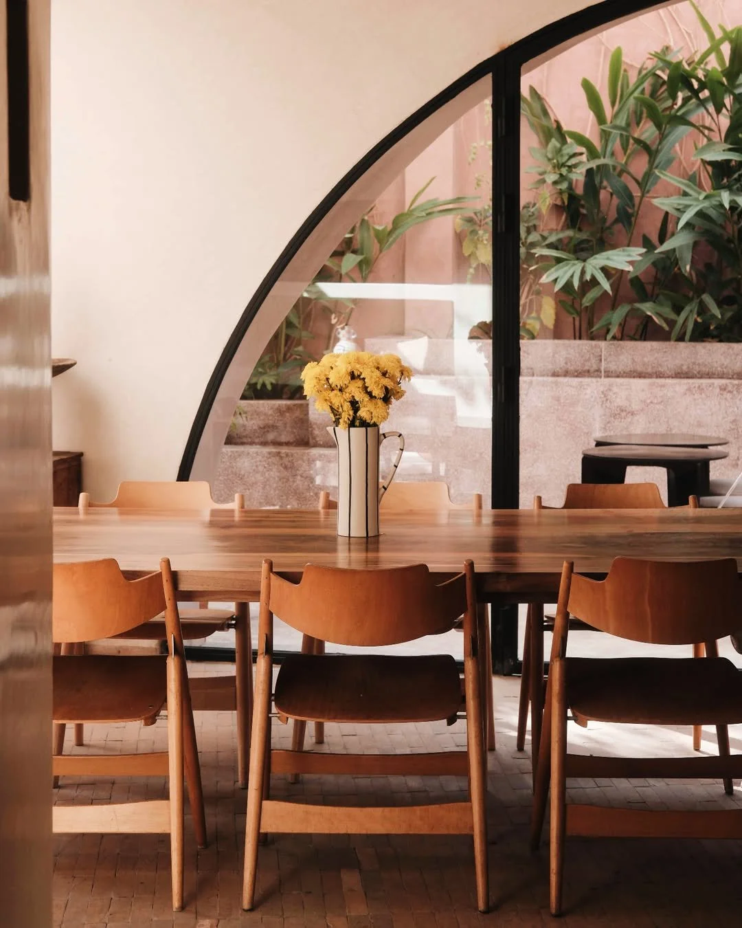 A wooden table decorated with a white vase filled with yellow flowers, set in a cozy indoor dining area with natural light, surrounded by wooden chairs and lush greenery outside a large window.