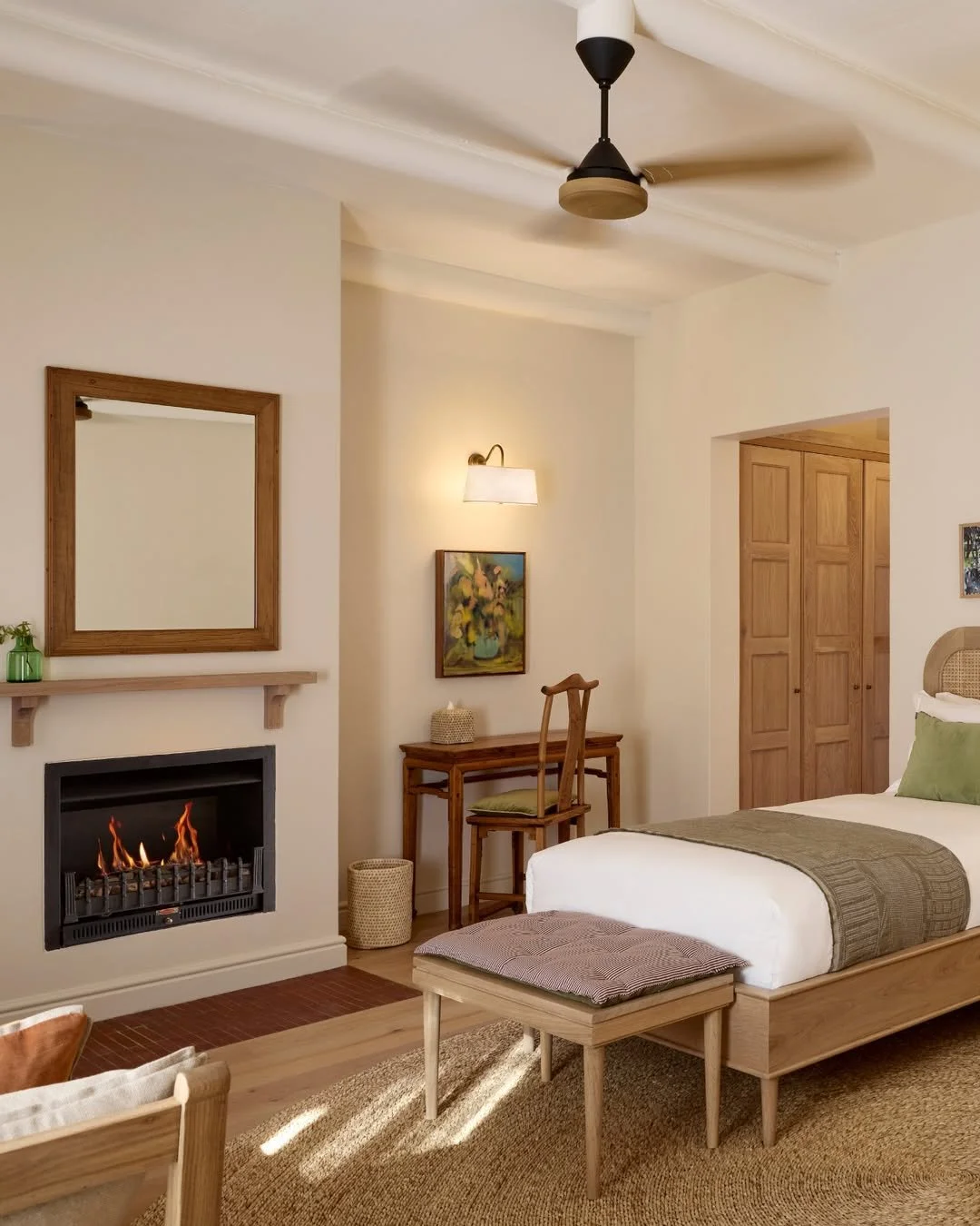 Hotel room with bed, fireplace, mirror, small desk, and wooden furniture, decorated in neutral tones.