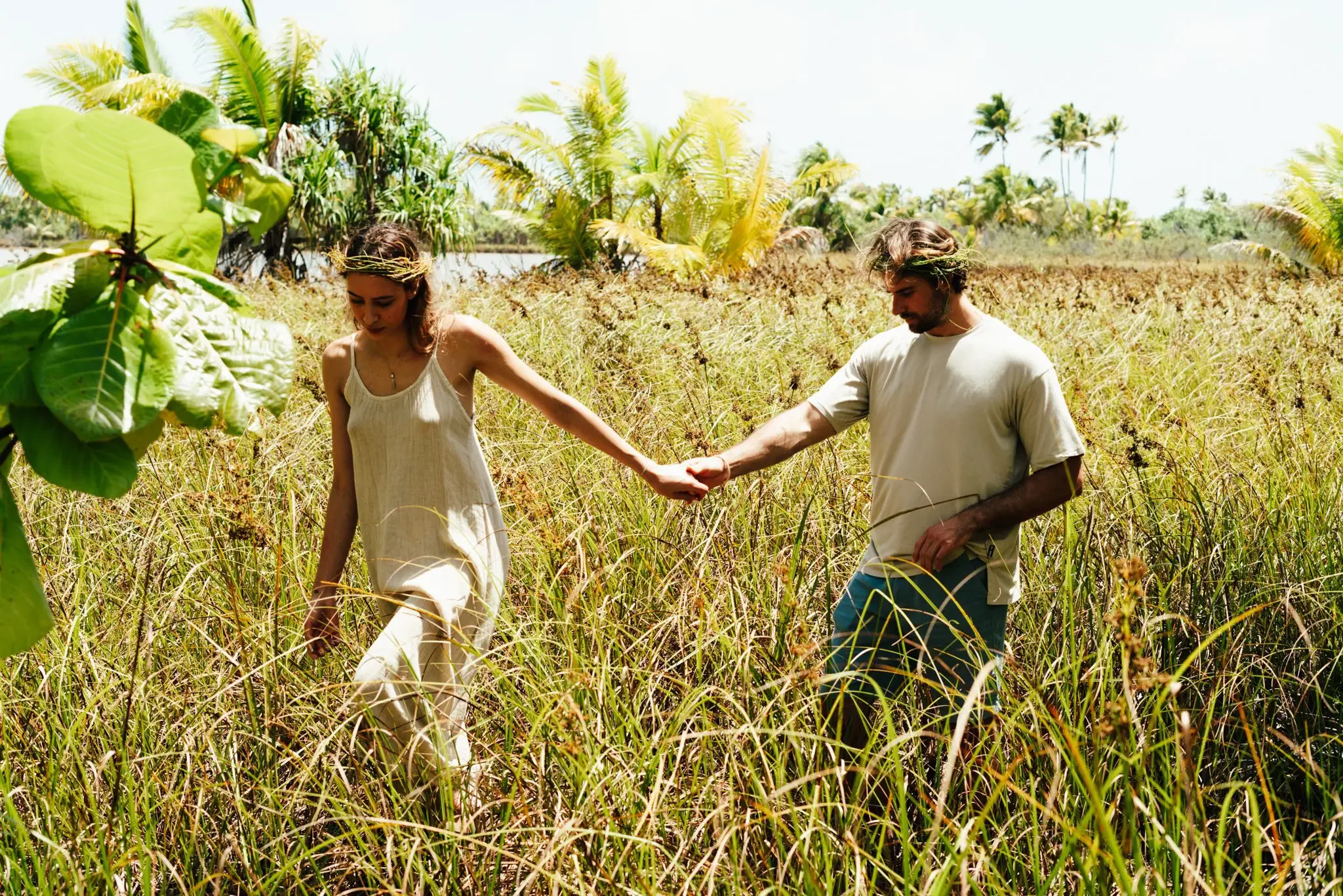 A man and woman holding hands and walking through a field of tall grass with palm trees and bright sky in the background, wearing casual clothing and plant crowns on their heads.