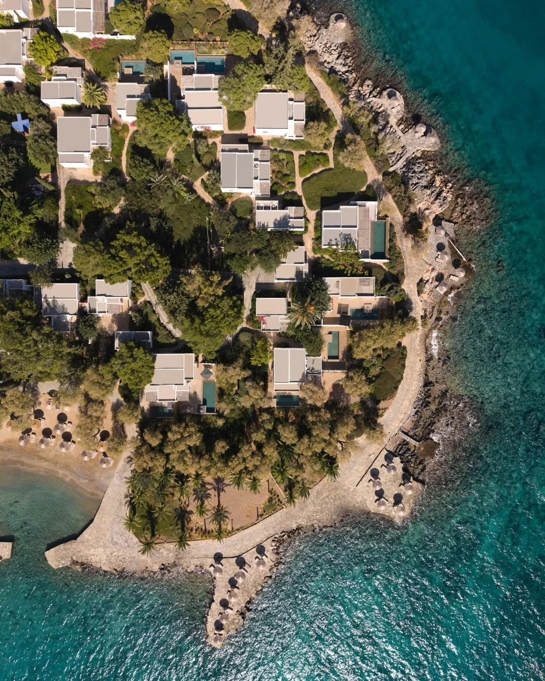 Aerial view of a coastal resort with white buildings, lush greenery, and pathways along a rocky shoreline with beach umbrellas and lounge chairs facing the ocean.