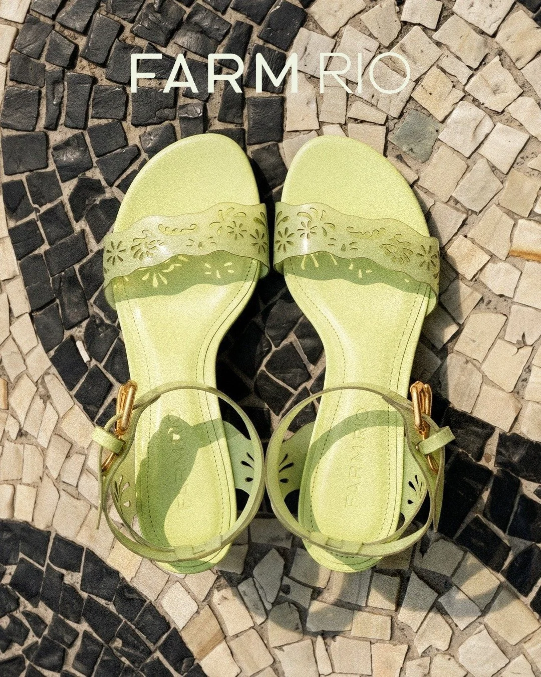 A pair of light green sandals with decorative cutouts and ankle straps with gold buckles, placed on a mosaic tile surface.