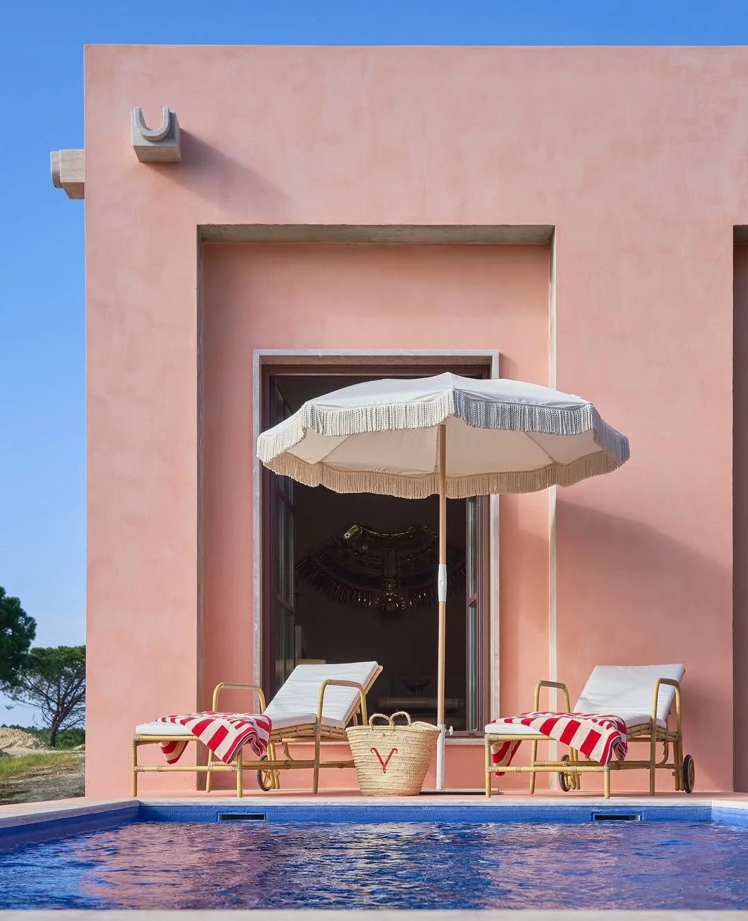Pink house with an outdoor swimming pool, two lounge chairs with red and white striped towels, a large white patio umbrella, a woven basket, and trees in the background.