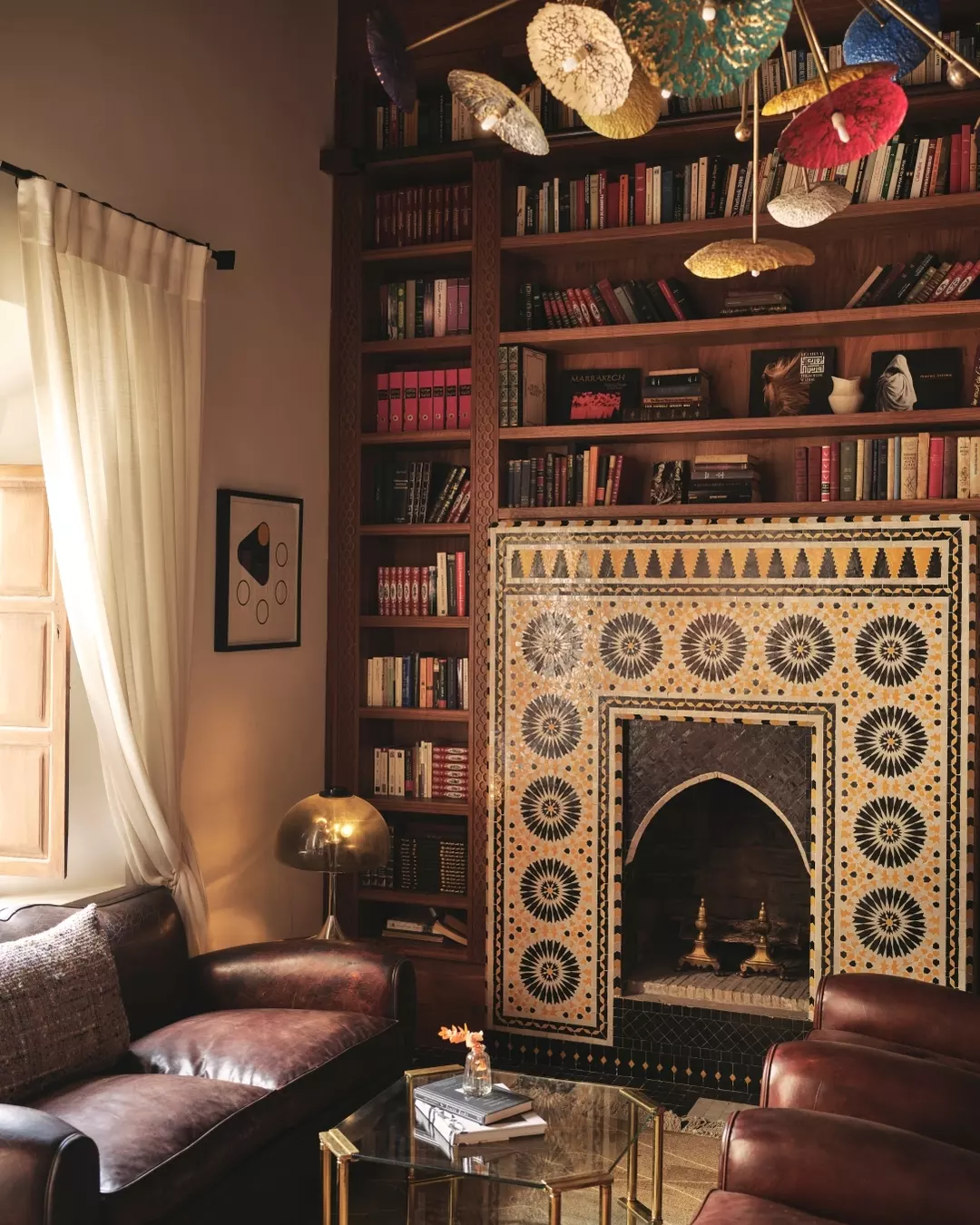 Cozy living room with a tall wooden bookshelf filled with books, a decorative fireplace with intricate tile work, a window with cream curtains, a black leather sofa, a brown leather armchair, a glass coffee table with books and a vase, a gold table lamp, and colorful mushroom-shaped pendant lights hanging from the ceiling.