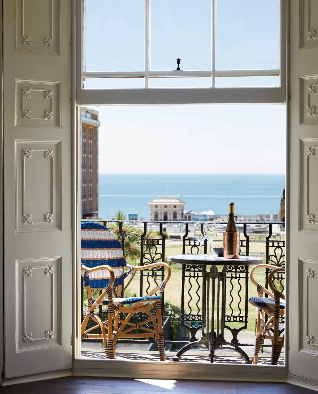 Balcony view with a small round table, two chairs, a bottle of wine, and two wine glasses overlooking the ocean and a historic building.