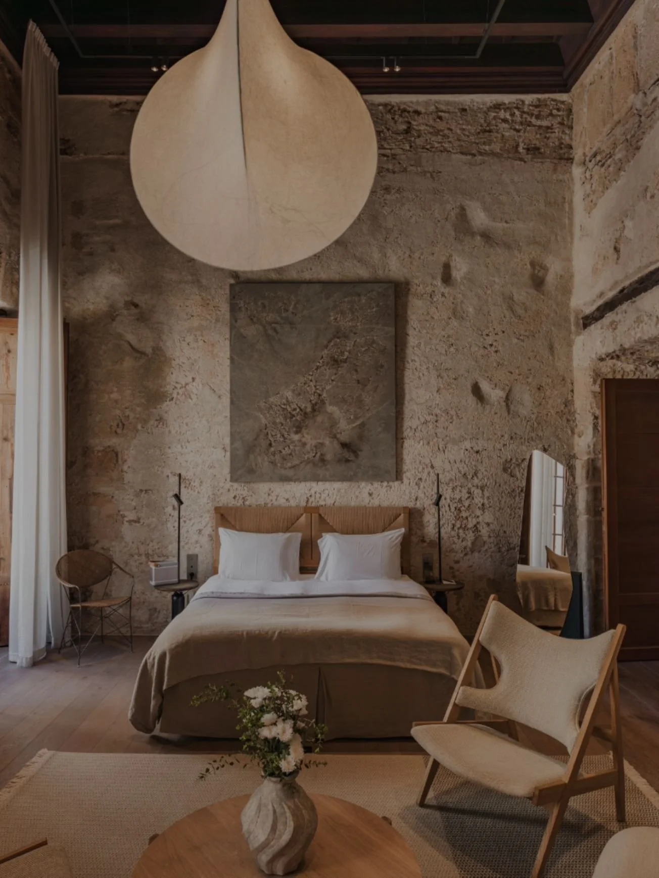 A rustic bedroom with exposed stone walls, a large bed with white pillows, a wooden headboard, and a large circular light fixture hanging from the ceiling. There are chairs, a side table, and a vase with flowers in the foreground.