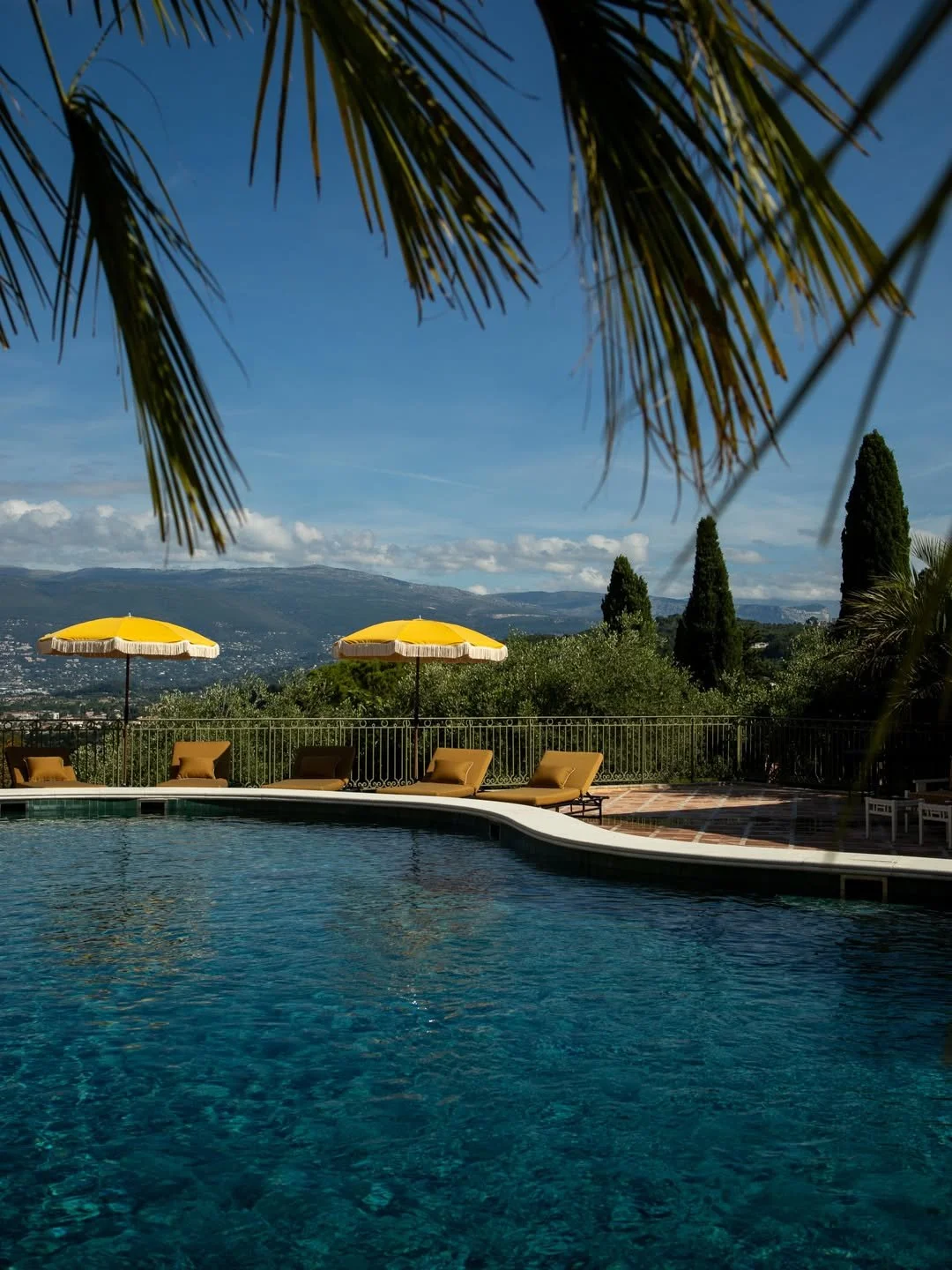 Swimming pool with yellow umbrellas and lounge chairs on a patio, overlooking a scenic view of trees, mountains, and a partly cloudy sky.