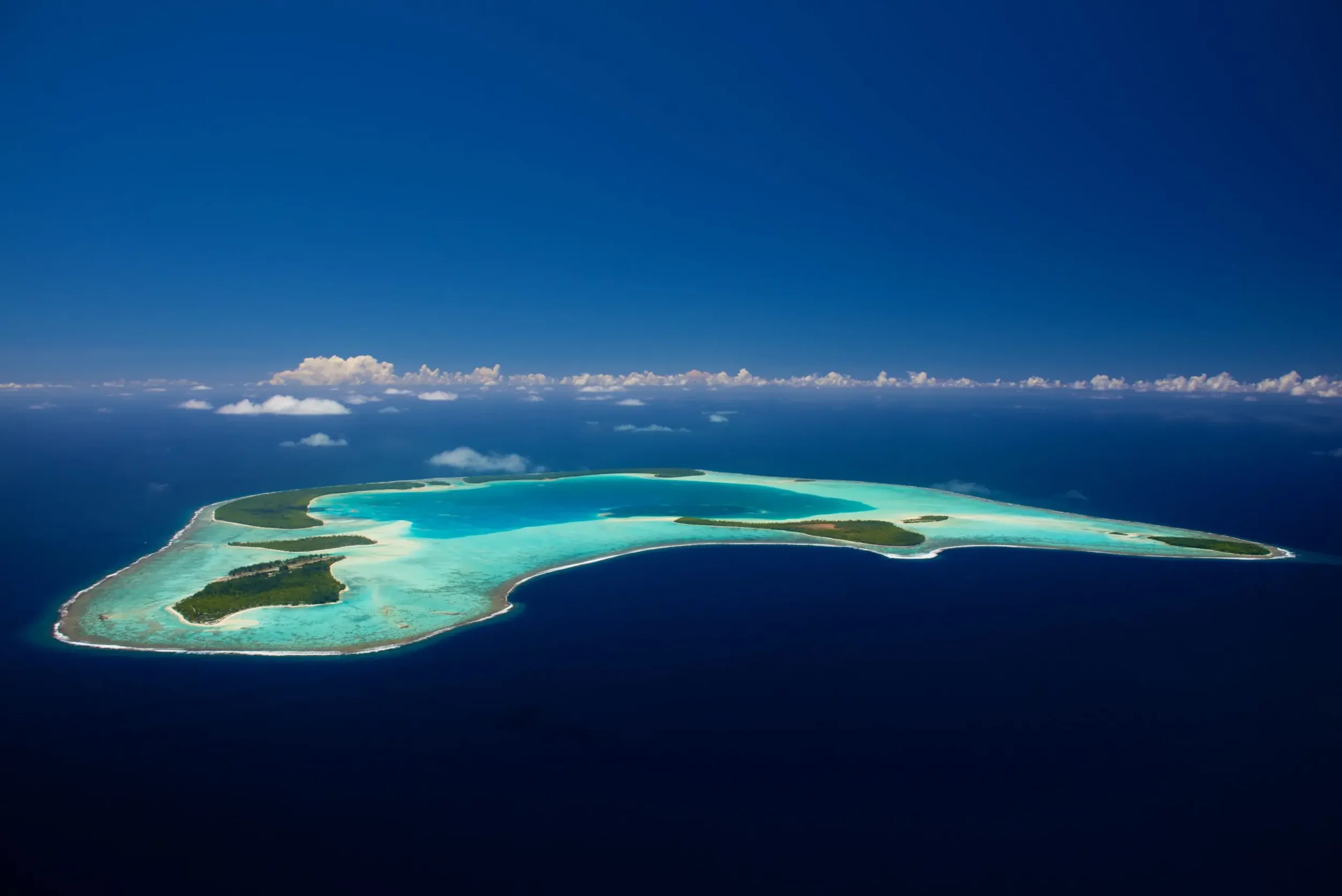 Aerial view of a tropical island with turquoise water surrounding it and a long sandy beach, under a clear blue sky with some clouds.