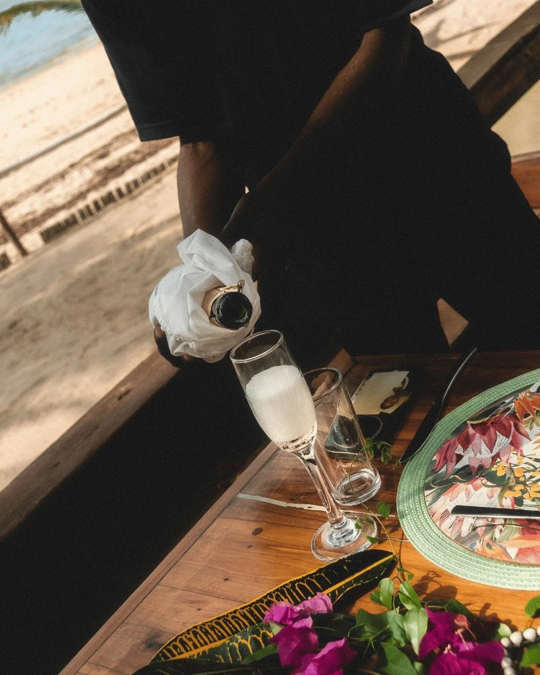 A person in black clothing is pouring champagne into a glass on a wooden table at a beachside setting before sunset.
