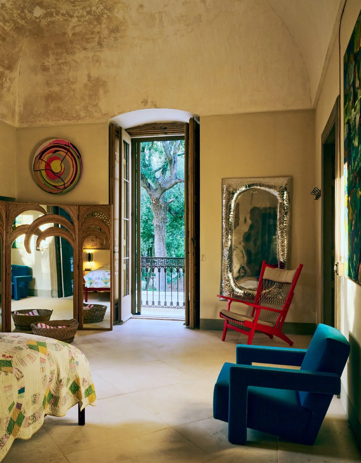 A colorful interior living space with open French doors leading to a balcony with trees outside. The room features various furniture pieces, including a red chair, a blue armchair, a large mirror with string lights, woven baskets, and a decorative partition. A painting hangs on the wall, and there is a bed with a quilted cover partially visible in the foreground.