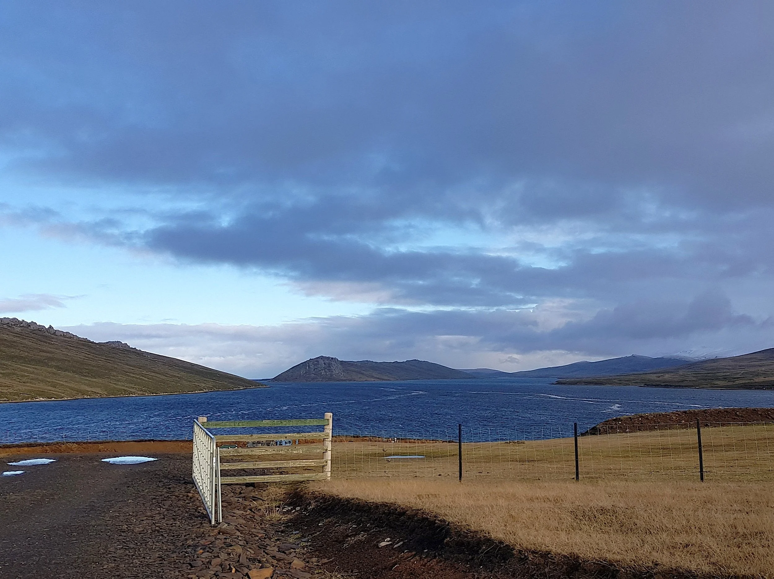 A landscape of a lake surrounded by hills and mountains under a partly cloudy sky with blue and gray clouds.